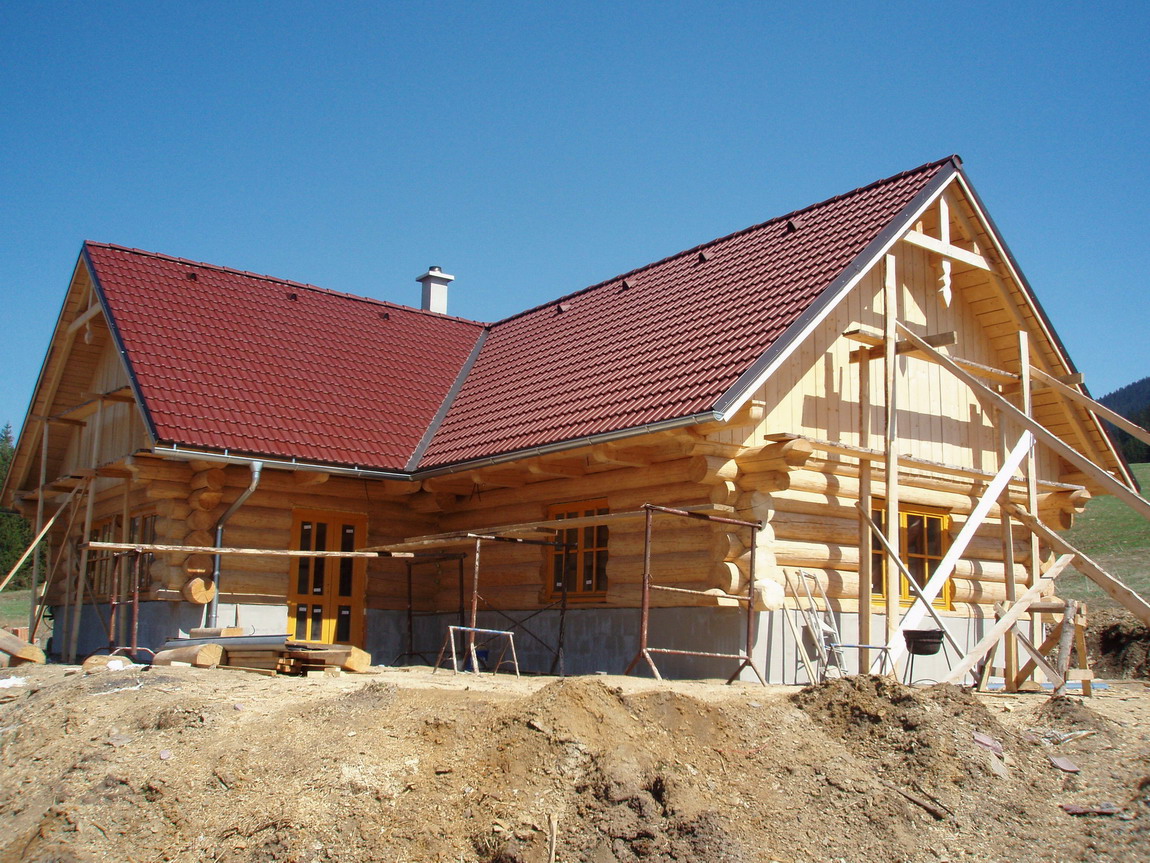 Surowy stan budowy domu z bali z czerwonym dachem, widoczne rusztowania i wykop ziemny, błękitne niebo w tle.